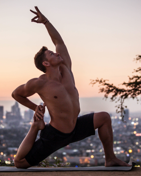 Coach Jake doing a yoga pose, reaching up to the sky with his left arm and extending his left knee forward and his right leg bent back, grabbing his right foot with his right hand with an urban backdrop