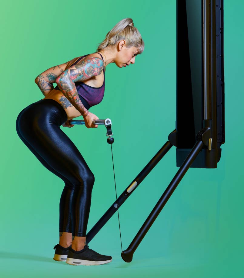 A woman in workout clothes performs a deadlift using her Tonal against a green backdrop.