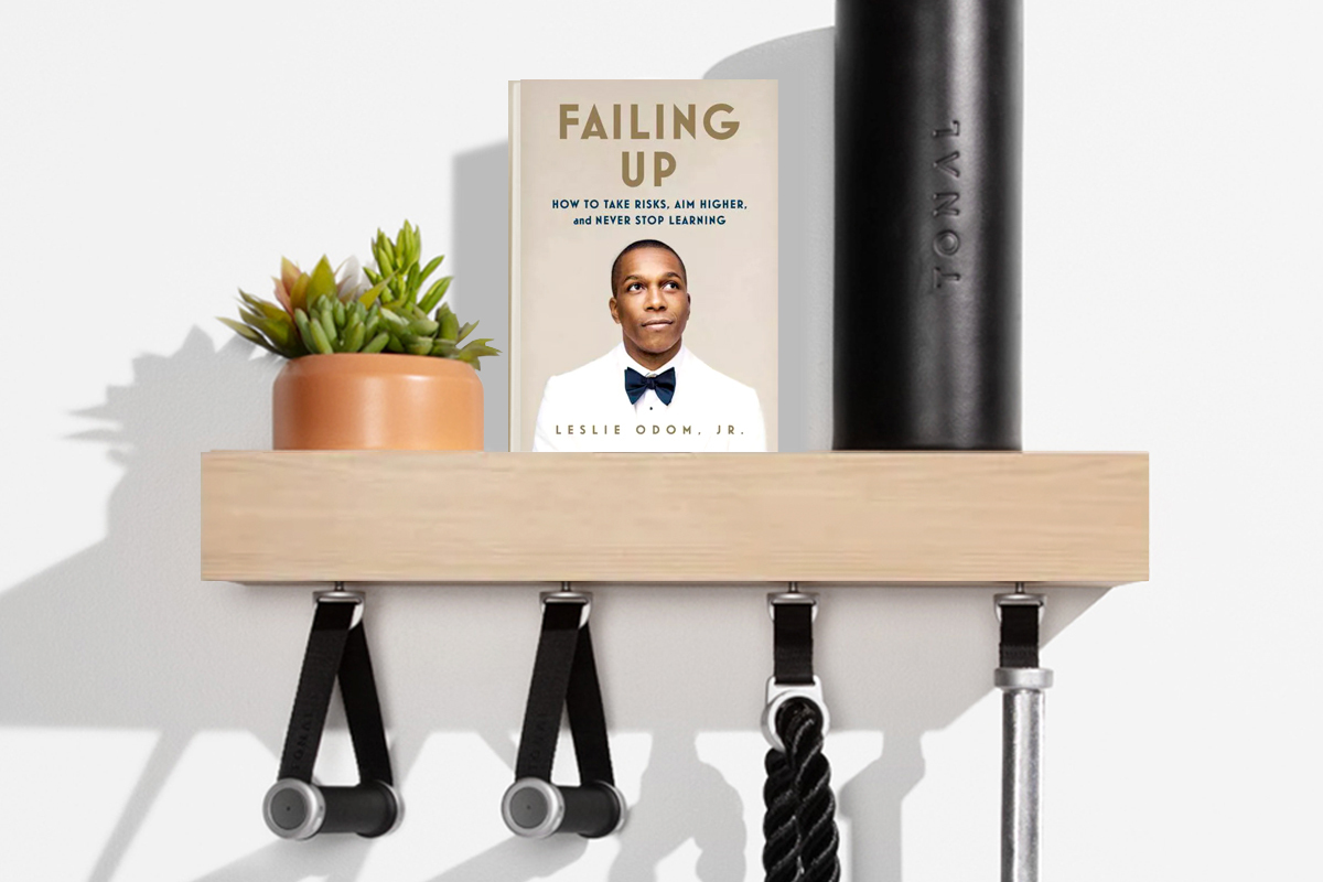 Failing Up book cover on a shelf next to a tonal foam roller and plant with weight lifting accessories hanging off it