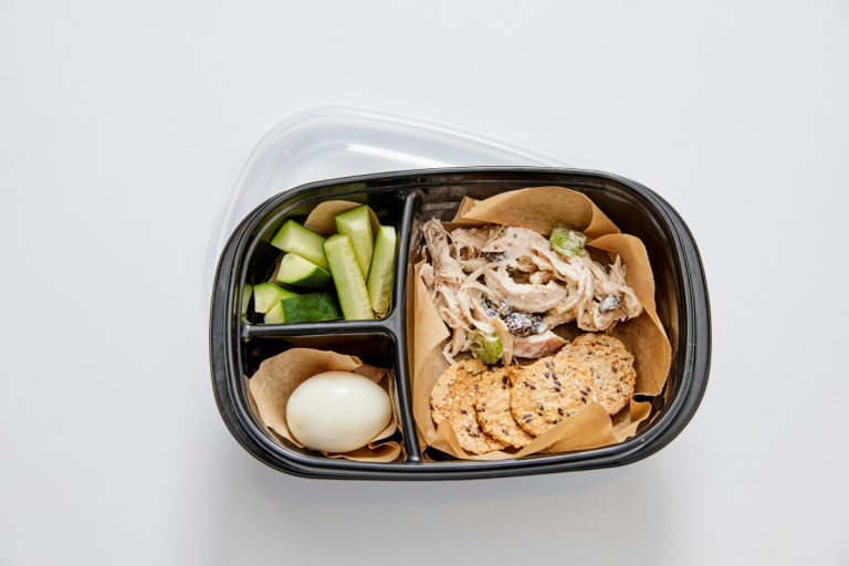 a container with chicken salad, an egg, crackers and some cucumbers